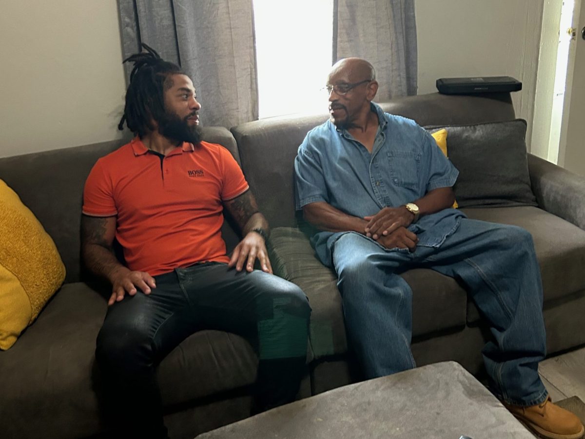 A man wearing an orange shirt and blue jeans sits on a sofa next to a man dressed in a denim shirt and pants. They are looking at each other and talking.