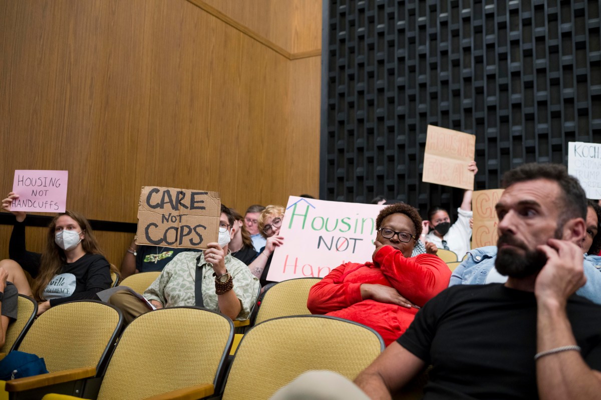 A photo inside Charlottesville City Council chambers, with people sitting in auditorium-style seats. A man in the foreground scratches his beard, while a woman behind him holds her head and smirks. In the background, people hold up signs that say "Housing Not Handcuffs," "Care Not Cops," and "Housing For All."
