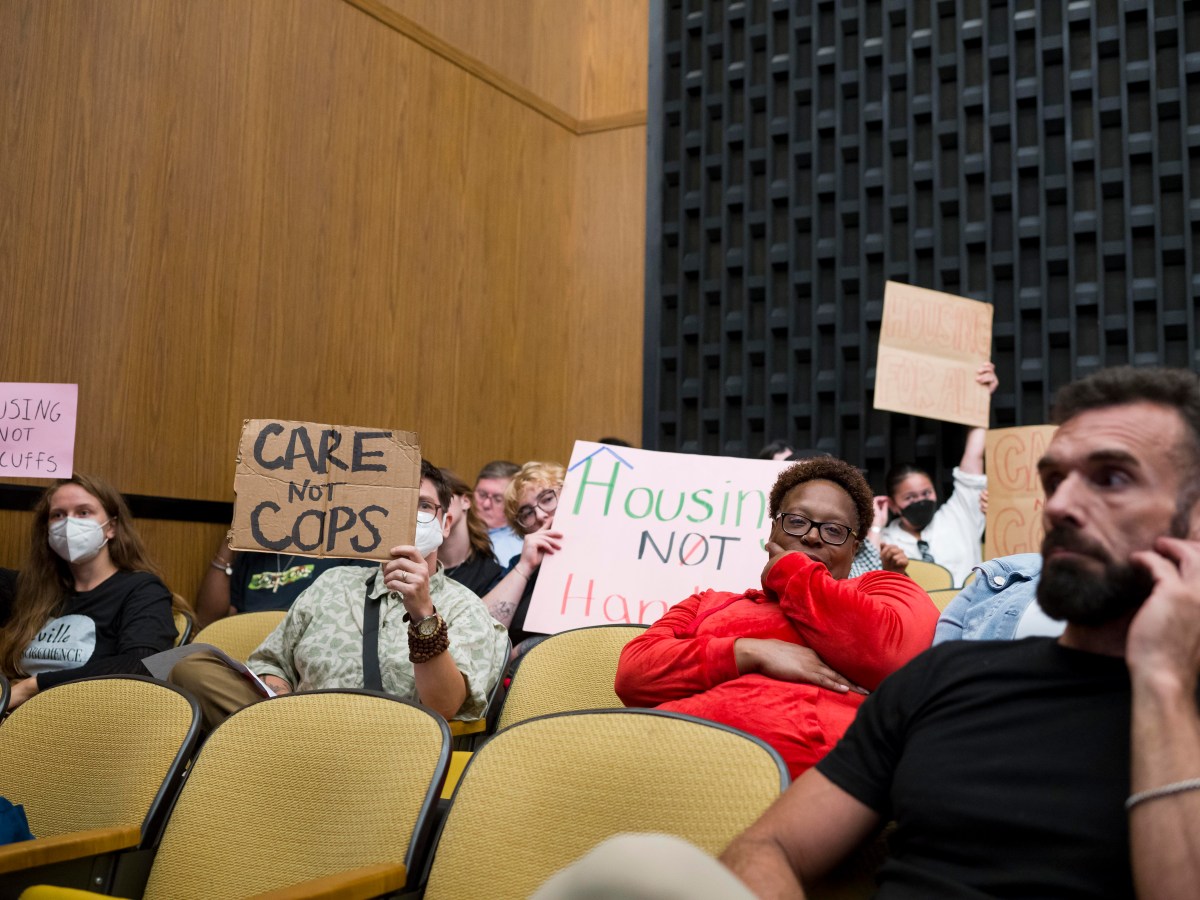 A photo inside Charlottesville City Council chambers, with people sitting in auditorium-style seats. A man in the foreground scratches his beard, while a woman behind him holds her head and smirks. In the background, people hold up signs that say "Housing Not Handcuffs," "Care Not Cops," and "Housing For All."