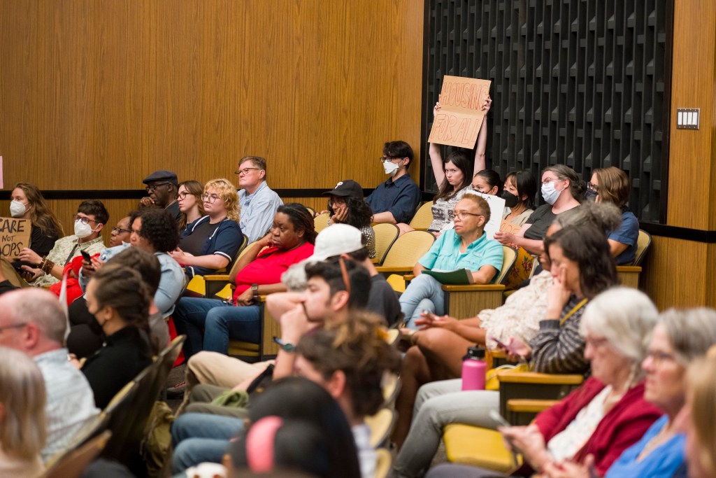 A view of Charlottesville City Council chambers, with most of the auditorium-style seats full. In the back corner, a young woman makes eye contact with the camera while holding up a sign that says "Housing For All."