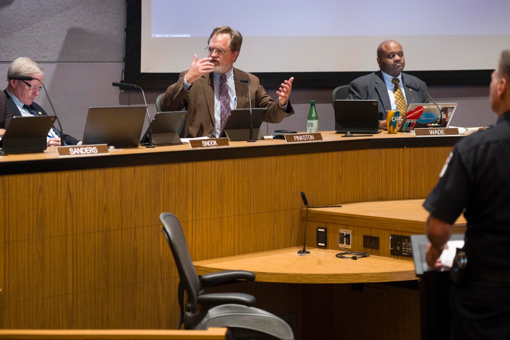 Charlottesville City Council chambers, with councilors seated at a long curved desk. The councilor to the left is looking down, presumably at the laptop and tablet in front of him, while the councilor in the middle is speaking and gesturing with his hands. The councilor to the right is looking out at the audience.