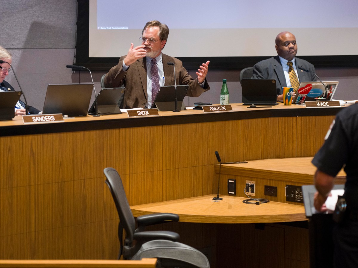 Charlottesville City Council chambers, with councilors seated at a long curved desk. The councilor to the left is looking down, presumably at the laptop and tablet in front of him, while the councilor in the middle is speaking and gesturing with his hands. The councilor to the right is looking out at the audience.