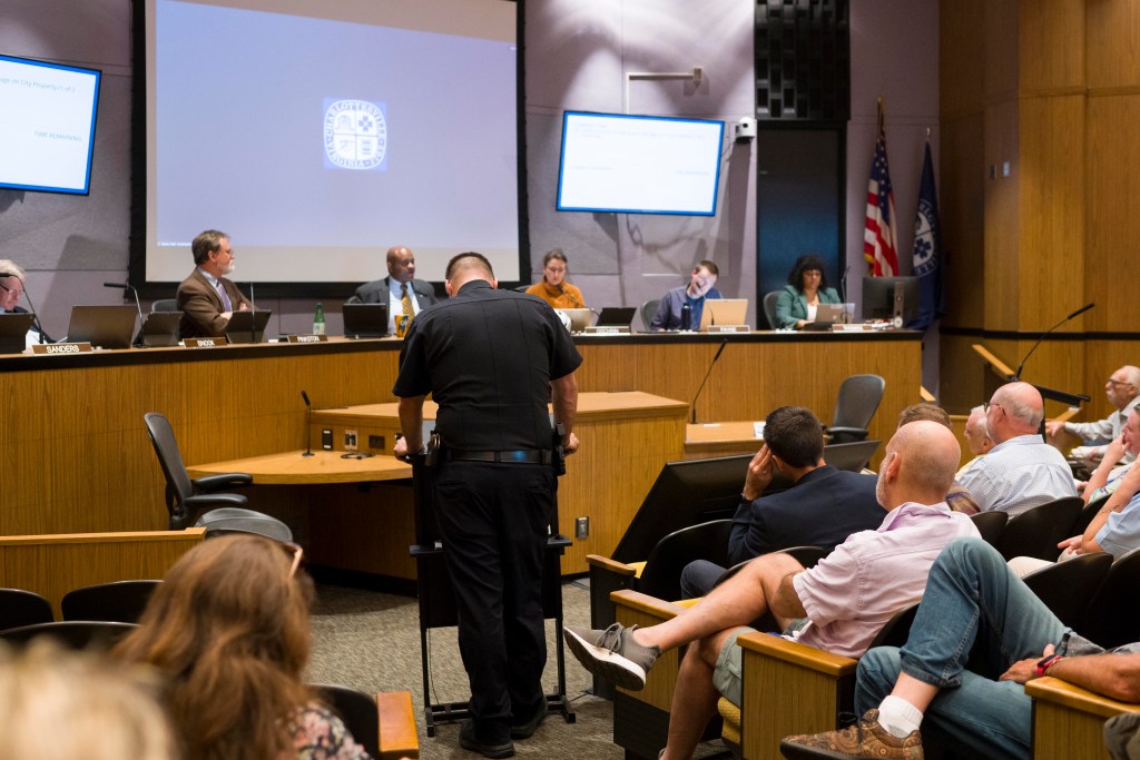 A man in a police uniform stands at a podium, facing a large curved desk at which five city councilors are seated, facing him and the large audience behind him.
