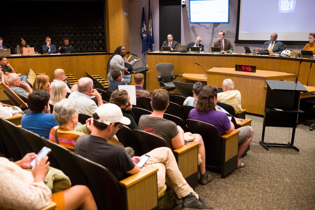 The inside of Charlottesville City Council chambers, with a young woman speaking at a podium, facing the audience. People sit in auditorium-style seating in the foreground, and city officials sit on a dais in the background.