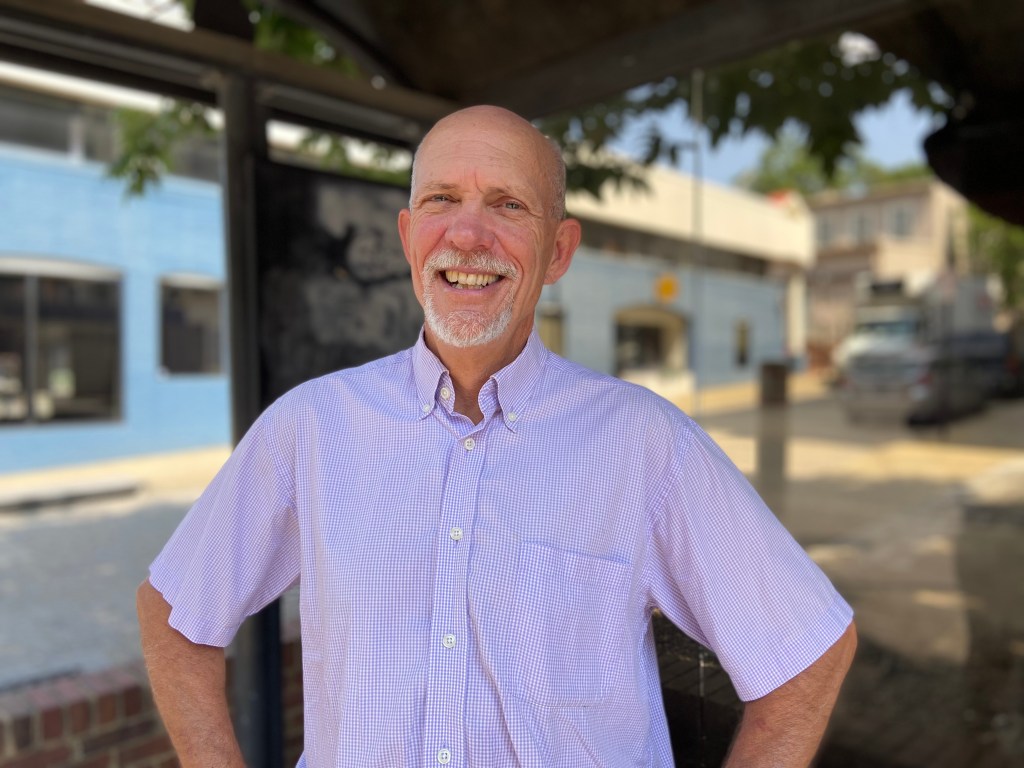 A person in a purple shirt stands at a bus stop, smiling.