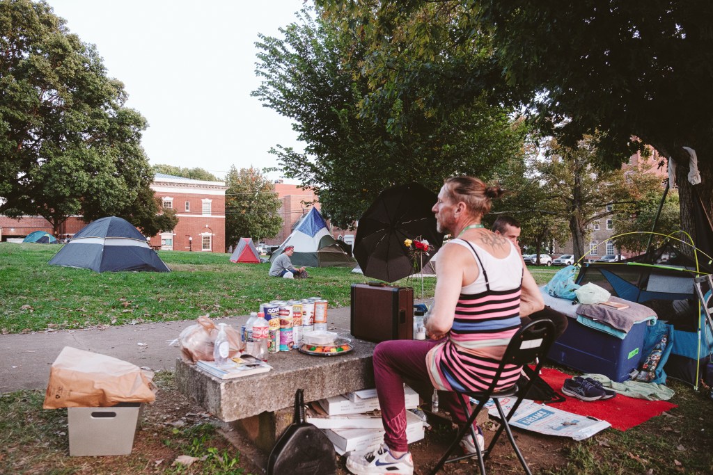 A photograph of a park in daylight. In the foreground, a person sits in a folding chair at a marble park bench. The bench is covered with canned goods, a plastic platter of food, and some empty plastic bottles. Nearby and in the background are multiple tents, and one person sitting on the grass.