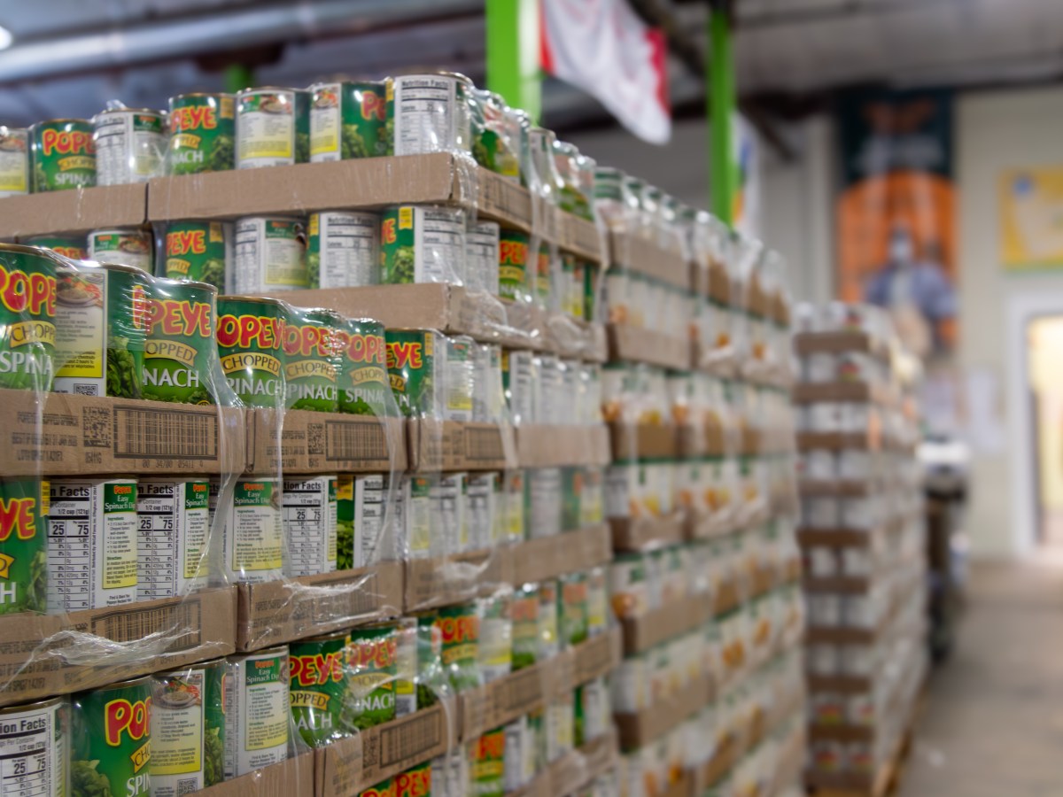 Wooden pallets stacked with large packages of canned spinach sit in a warehouse. An employee in a bright orange shirt is visible in the background.