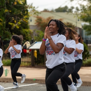 A group of young women wearing matching shirts that read "cheer" in large letters clap their hands in unison.