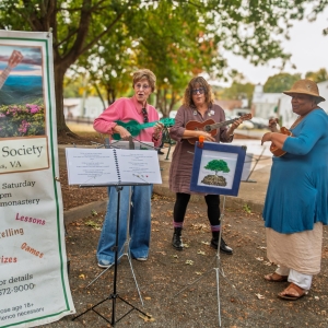 Three women stand under trees singing and playing ukuleles. They have music stands in front of them and a large sign next to them reading "Ukulele Society, Nasons, VA"