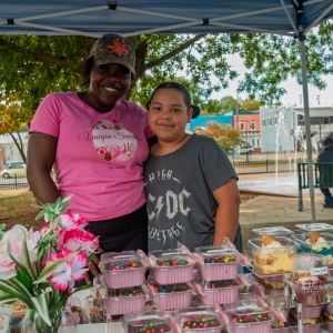 Two women stand under a large canopy with their arms around each other, smiling at the camera. On a large table in front of them is a a vase with flowers and several different types of candies and cakes.