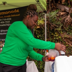 A woman stands under a canopy mixing ingredients into a large plastic cup full of ice and lemonade. A large sign behind her reads "Lay's Lemonade."