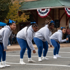 A group of young women wearing matching white socks, blue pants and grey sweatshirts dance on a street in front of a large brick building with a green awning decorated with flags. A small crowd of people in the background watches them perform.