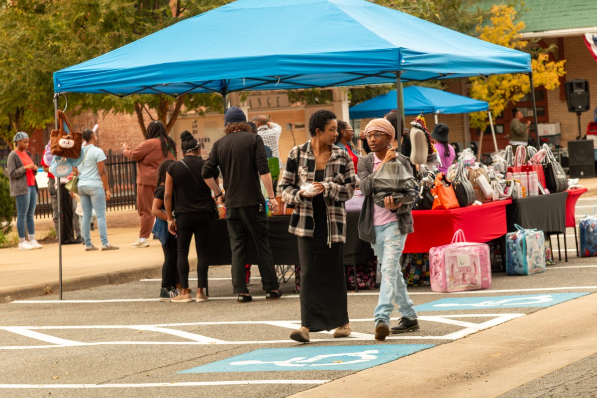 Two people walk across parking spaces next to a street. Large canopies, tables and crowds of people are visible behind them.