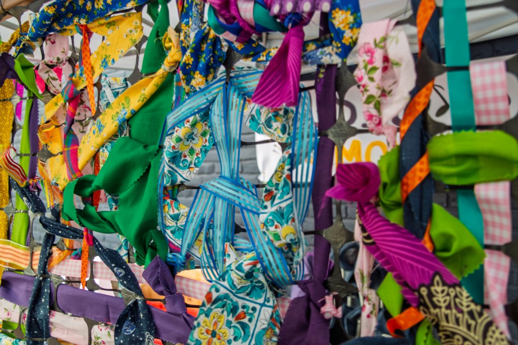 Several strips of colorful cloth are weaved between the netting of a safety fence, making various designs, including a large peace symbol in the center.