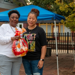 Two women stand close to each other holding a colorful basket wrapped in plastic. A brick sidewalk with trees and a small canopy are visible behind them.