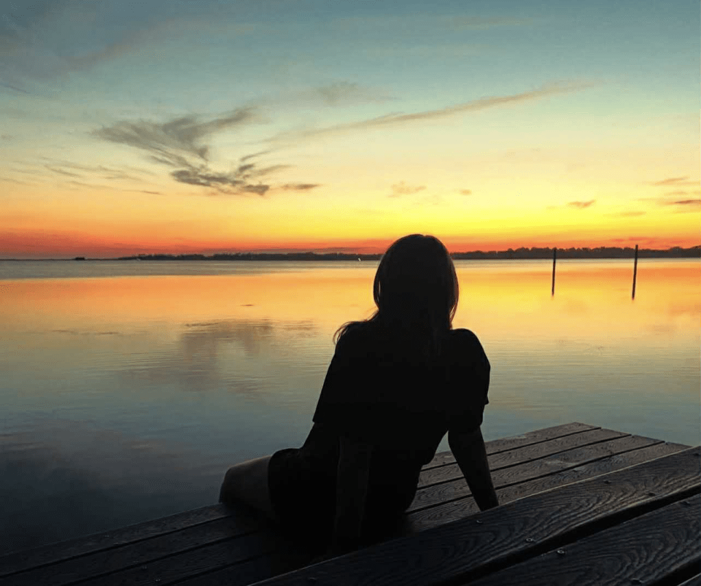 A dark silhouette of a woman sitting on a deck and watching a sunset over a large body of water.