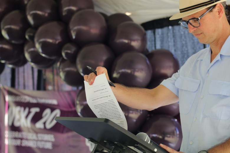 A man reads notes at a podium. Behind him there is a balloon arch.