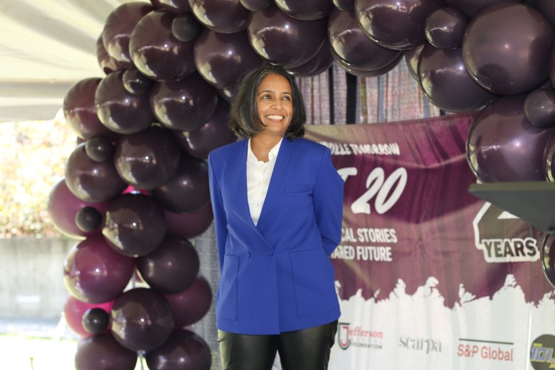 A woman smiles. She stands in front of a balloon arch and a banner that says NEXT 20.