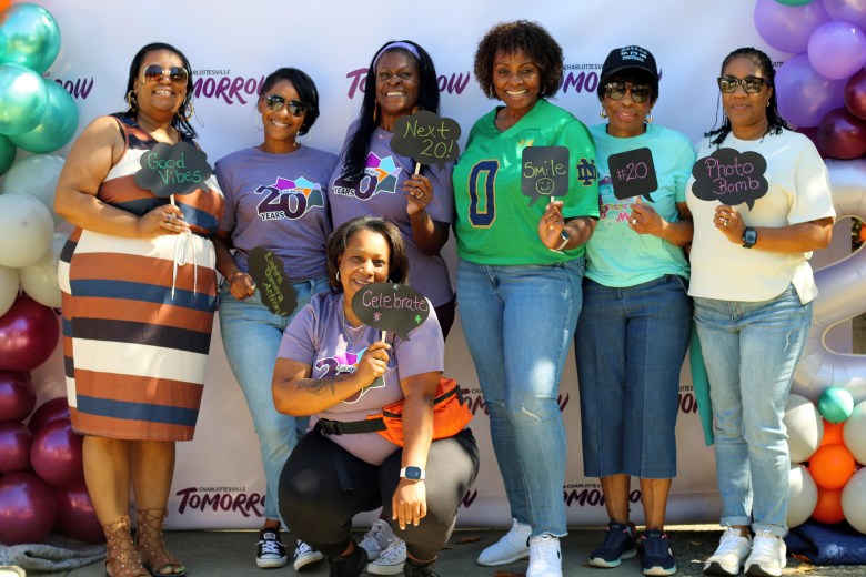 A group smiles, holding signs that say GOOD VIBES, CELEBRATE, NEXT 20, SMILE, #20, and PHOTO BOMB. They pose in front of a backdrop that says CHARLOTTESVILLE TOMORROW.