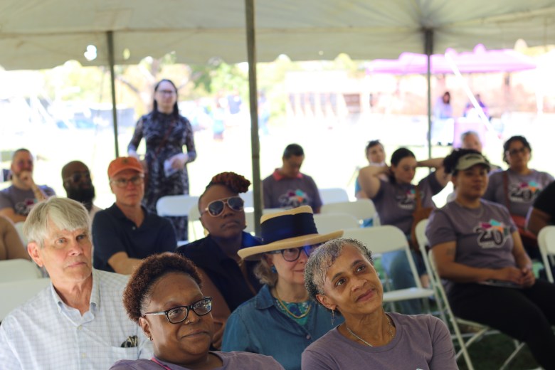 An audience sits in folding chairs under a tent.