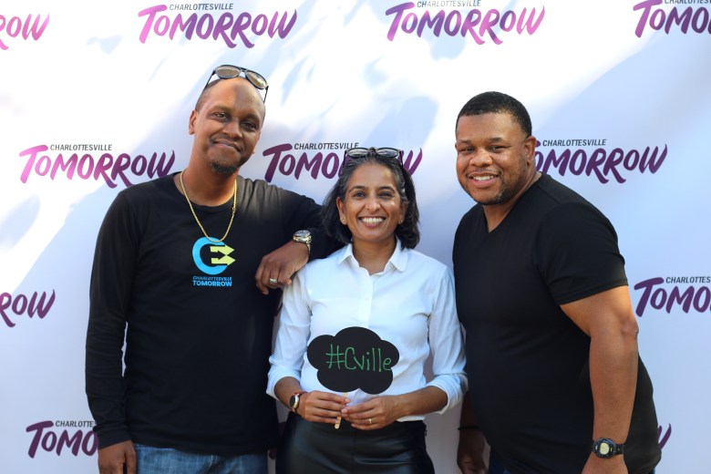 Three people smile. One holds a sign that says #CVILLE. They stand in front of a backdrop that says CHARLOTTESVILLE TOMORROW.