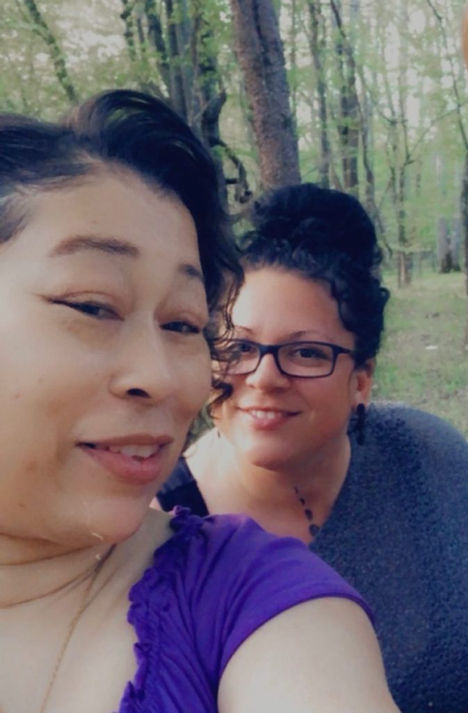 Two women look at the camera, smiling, with trees visible behind them.