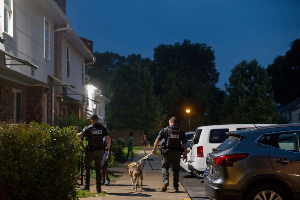 Two men in tactical gear and bulletproof vests walk down a sidewalk in a residential area at night with a German Shepard dog.