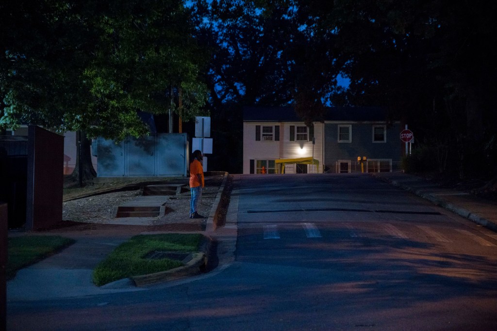 A man in a bright orange shirt stands at a residential intersection at night. The neighborhood is empty apart from him.