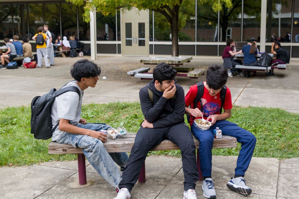 Three high school students wearing backpacks sit on a bench in a courtyard, talking and eating lunch.