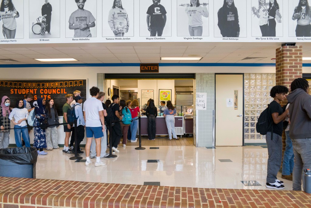 Several high school students line up next to a white wall with a large bulletin board. The line leads into a doorway where cafeteria workers are visible serving lunches.