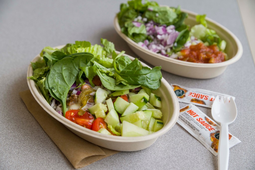 Two disposable bowls filled with rice and various vegetables on a table, with napkins, a white plastic spork and small packets of hot sauce sitting beside them.