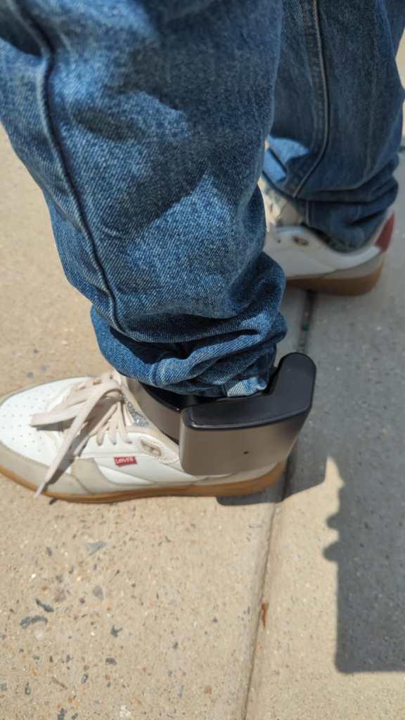 A close up photo of an ankle monitor around the ankle of a person wearing jeans and white shoes.