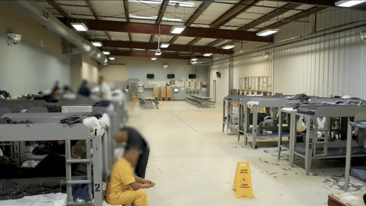A large open room with several bunk beds lined up against two walls and tables visible in the distance. People in jumpsuits stand or sit near the bunk beds.