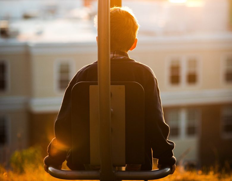 A young boy sits on a swing, his back to camera, and watches the city at sunset.