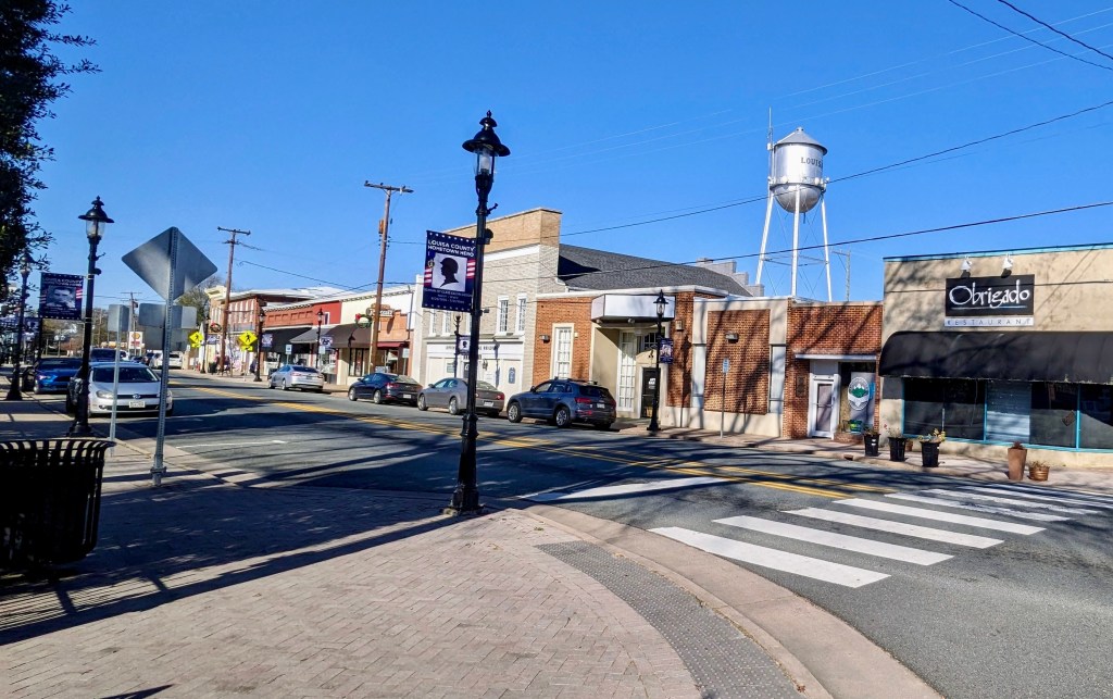 A small-town main street with shops, parked cars, streetlights and a water tower under a clear blue sky.