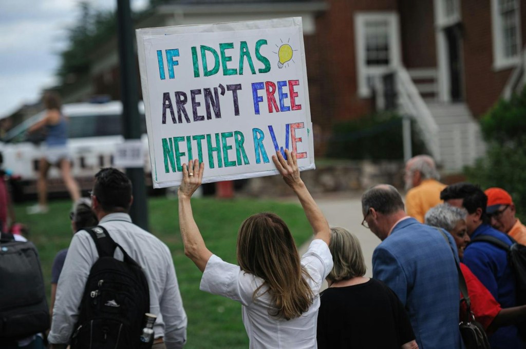 A crowd of people gather on a lawn next to large brick buildings with white trim. One person is holding up a large sign that reads "If ideas aren't free neither are we"