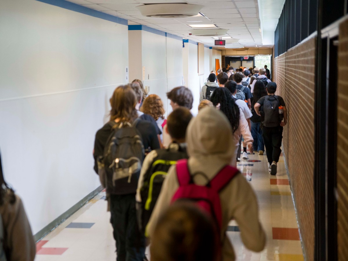 A view of an indoor hallway with a white wall on the left, a brick wall on the right, and colorful tiles on the floor. Several high school students wearing backpacks walk down the hallway, with their backs facing the camera, and a crowd of students is visible at an intersecting hallway in the distance.