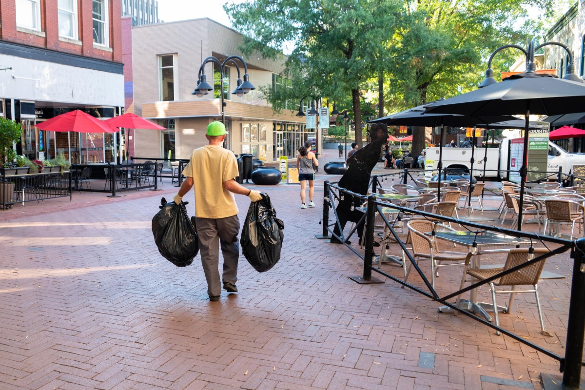 A photo of an outdoor pedestrian mall paved with bricks. A man in a t-shirt, work pants, a baseball cap and work gloves carries two plastic trash bags toward a white truck in the background. The truck is partially obscured by umbrellas from a restaurant's outdoor patio, a map of the pedestrian mall and directional signs for businesses.