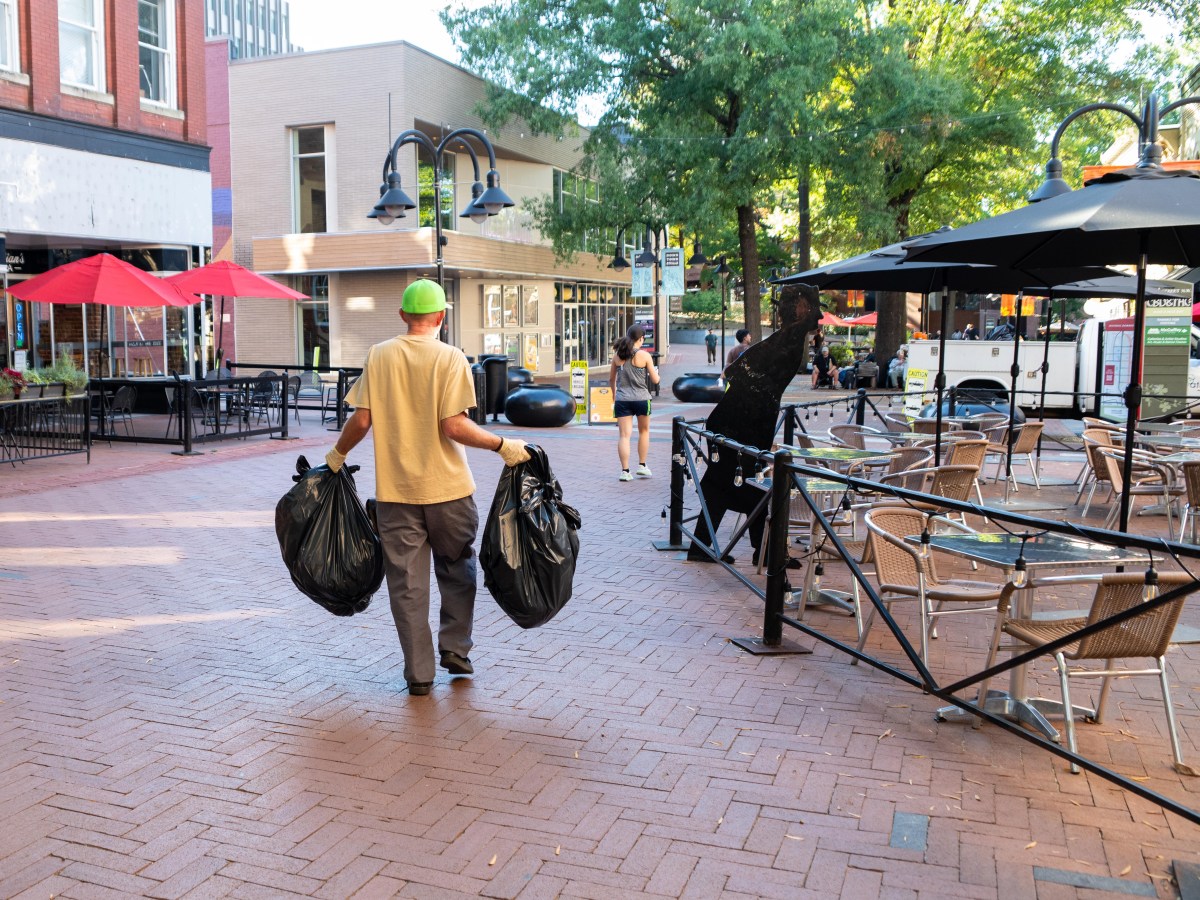 A photo of an outdoor pedestrian mall paved with bricks. A man in a t-shirt, work pants, a baseball cap and work gloves carries two plastic trash bags toward a white truck in the background. The truck is partially obscured by umbrellas from a restaurant's outdoor patio, a map of the pedestrian mall and directional signs for businesses.