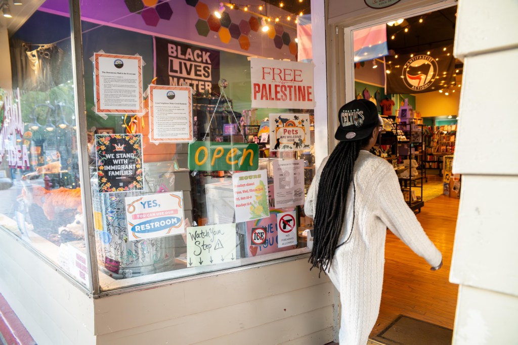 A person wearing a long cable-knit sweater and a Boston Celtics baseball cap steps into a shop. The shop windows are full of signs that read "we stand with immigrant families," "yes, you can use our restroom," "watch your step," "free Palestine" and "pets are welcome here."