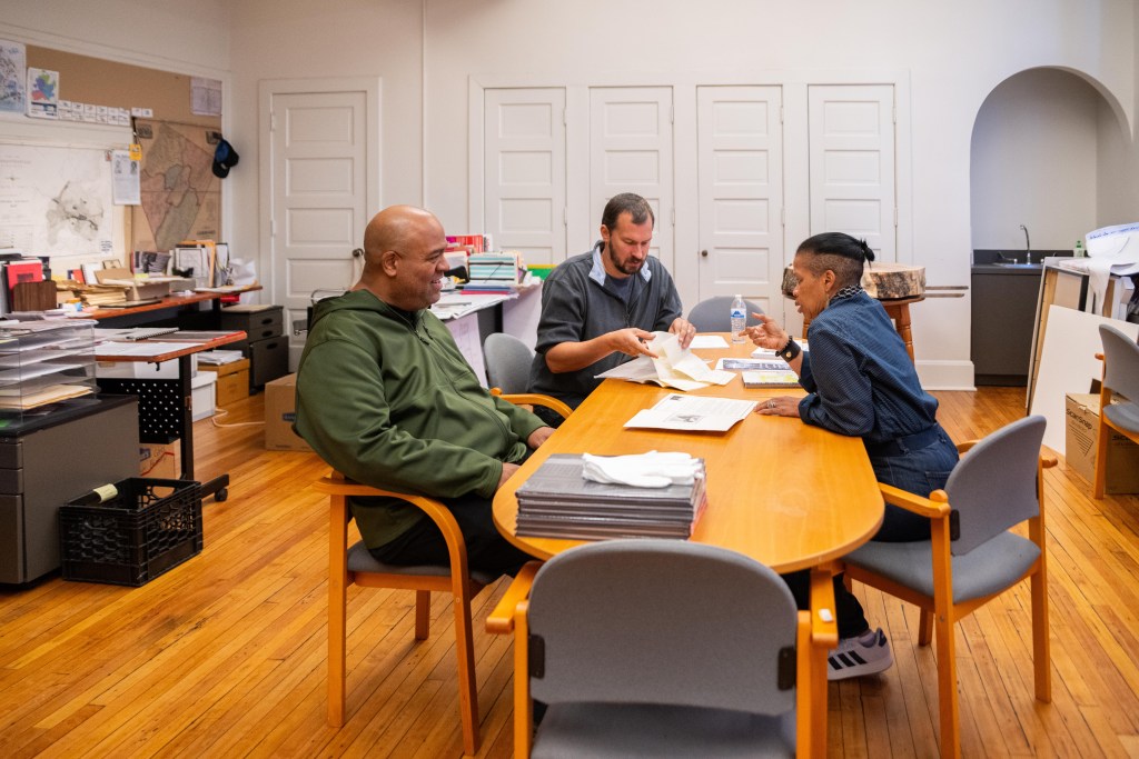 Two men and a woman having a conversation while sitting around an oval-shaped table. There are piles of papers and documents on the table, and one of the men is sifting through them. They're in an old classroom with wood floors. The walls are lined with maps and desks piled high with manila envelopes and boxes full of documents.