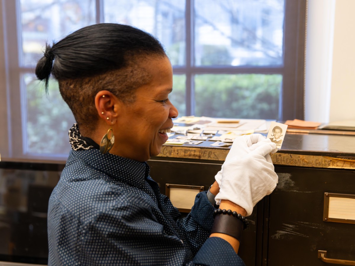 A photograph of a woman wearing white gloves holds a small school photo of a young girl. The woman is smiling wide as she affectionately points to the girl's face. The woman is standing in front of two tall filing cabinets, and there are more photos on top of the cabinets.