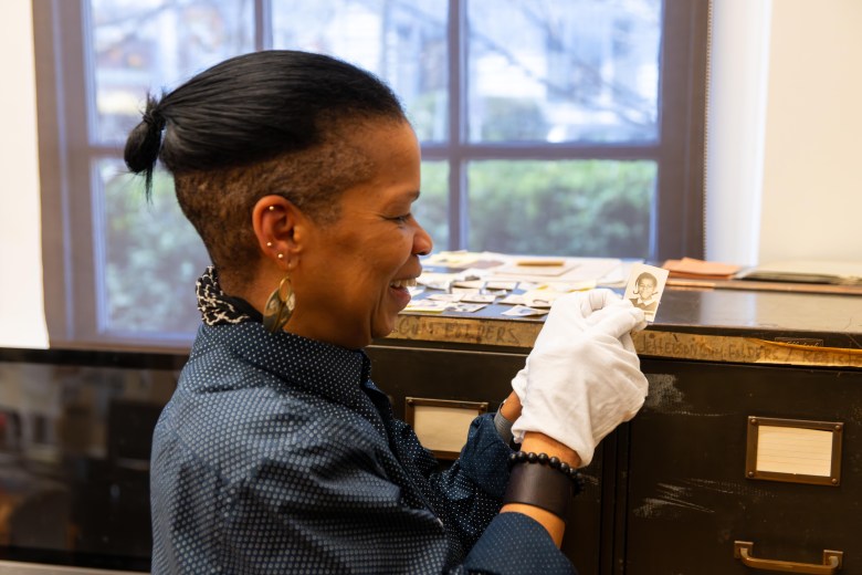 A photograph of a woman wearing white gloves holds a small school photo of a young girl. The woman is smiling wide as she affectionately points to the girl's face. The woman is standing in front of two tall filing cabinets, and there are more photos on top of the cabinets.