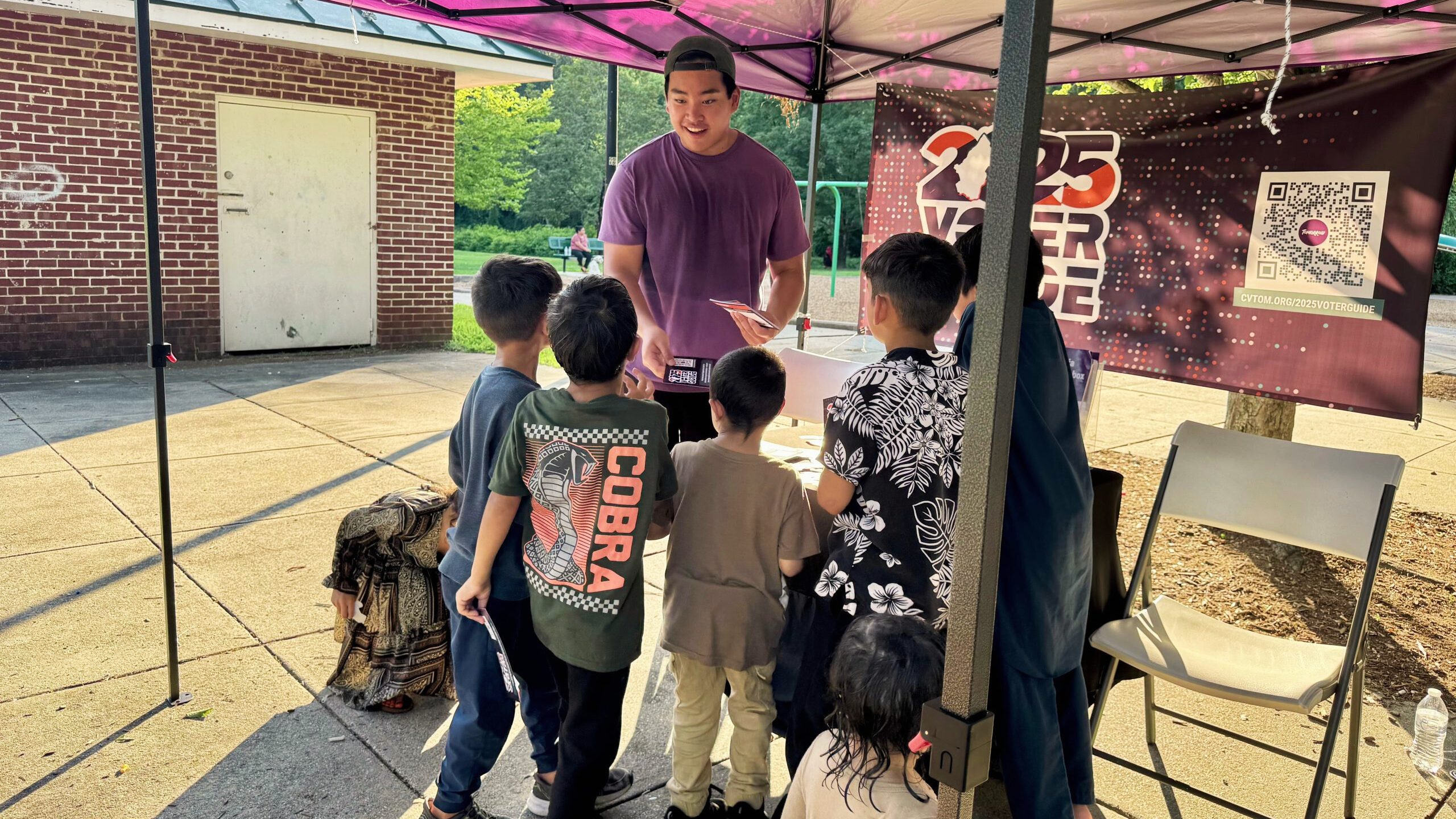 A young man interacts with a group of children under a tent at an outdoor event. The tent has a banner promoting the 2025 Voter Guide. The setting appears bright and lively, with a playground visible in the background.