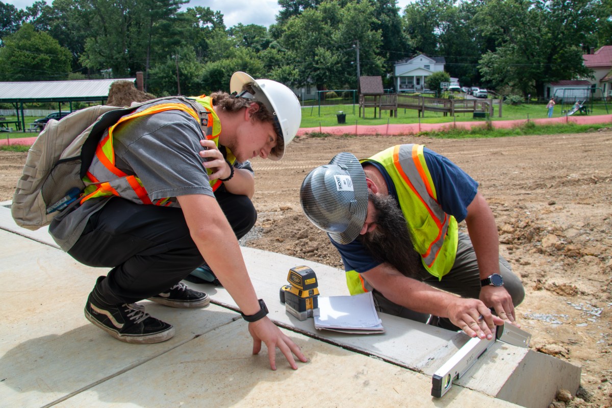 Two people in hard hats and safety vests measure an area on a cement slab.
