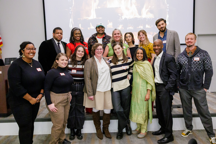 A group of people standing in two rows posing for the camera, with a projected image of a fireplace behind them.
