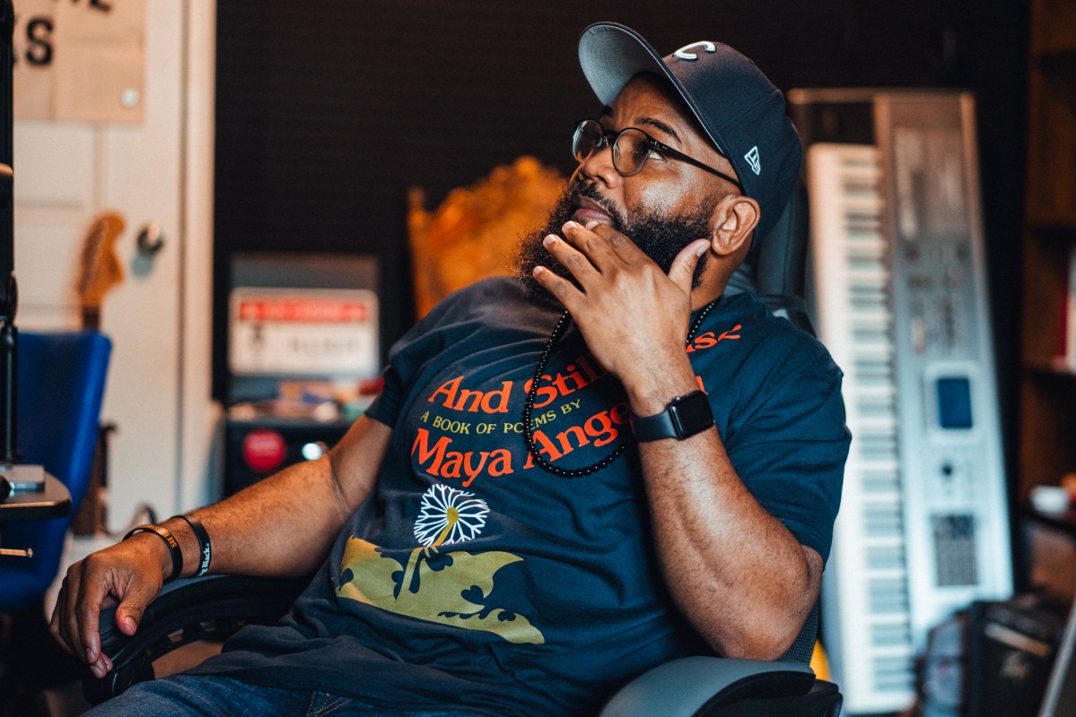 A man wearing glasses and a baseball cap reclines in a chair next to a desk, holding his hand up to his chin. A chest of drawers and various musical instruments, including an electric guitar and a keyboard, are visible behind him.
