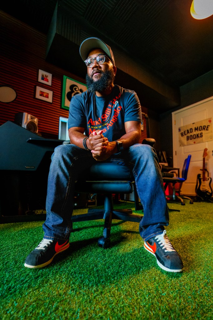 A man wearing a baseball cap, blue T-shirt and denim jeans sits in a chair in his office. An electric guitar and a sign reading "read more books" are visible behind him.