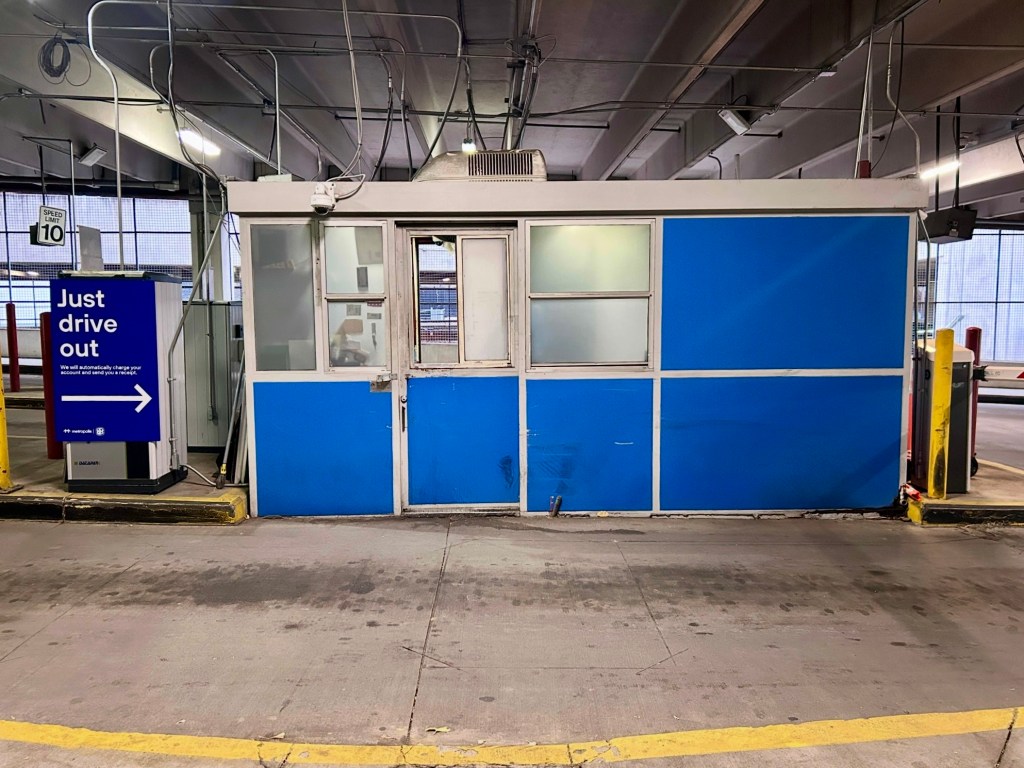 A photograph of the inside of a parking garage. In the center of the photo is a booth where a garage attendant typically sits during daytime hours. To the left of the booth is a sign that reads "just drive out. We will automatically charge your account and send you a receipt," with an arrow pointing toward the exit.