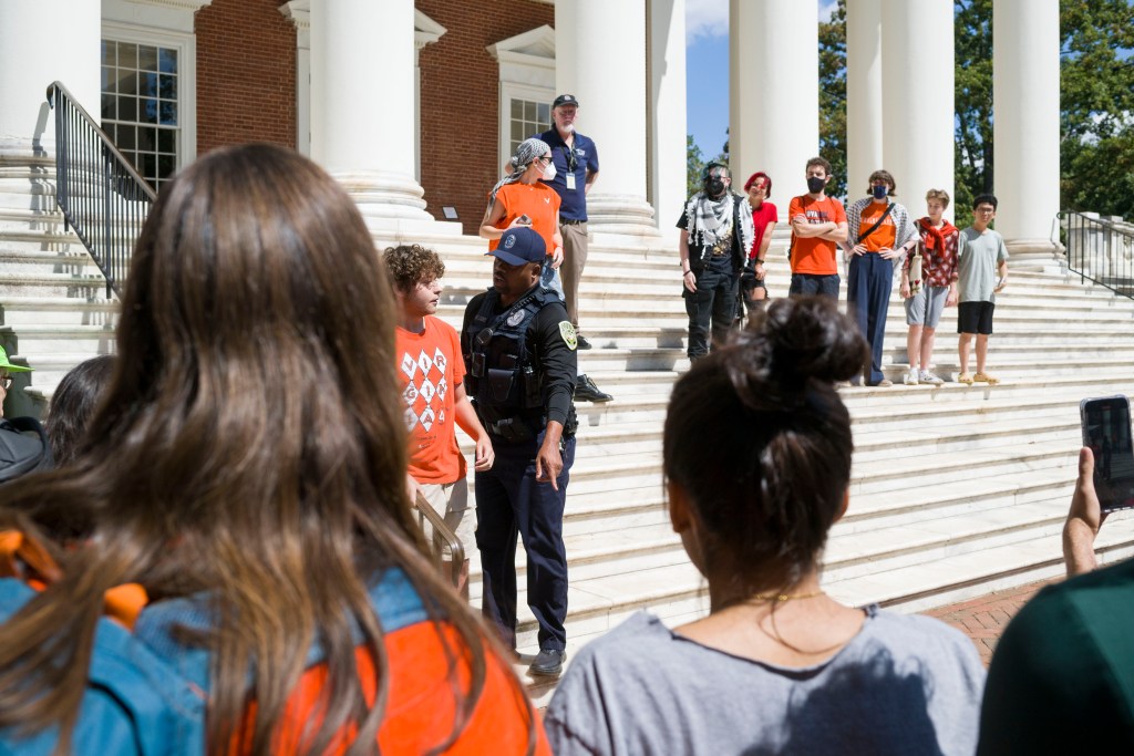 A police officer wearing a kevlar vest and blue baseball cap points down while addressing a man in an orange shirt as they both stand on the wide white steps of a large brick building with white columns. A row of people observing them — some also wearing orange shirts as well as face masks — stand behind them and to the right.